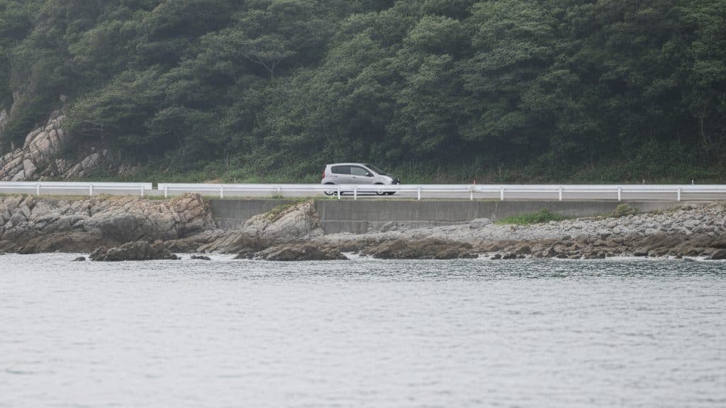 岩場の海岸に沿って続く道を、海の向こうから見た写真。白いガードレールの道を銀色の小さな車が横向きに走り、道の後ろには木々に覆われた斜面が迫り、手前には穏やかな海が広がっている