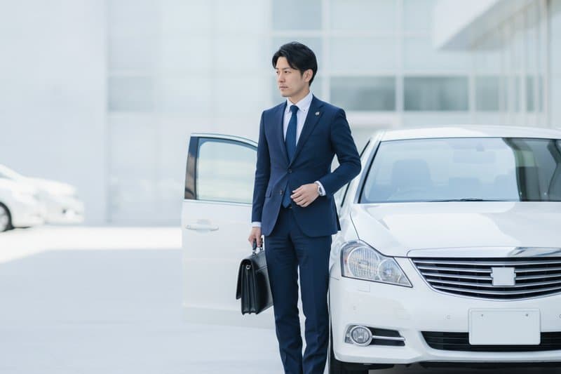 白い営業車の横に立つスーツ姿の営業担当者